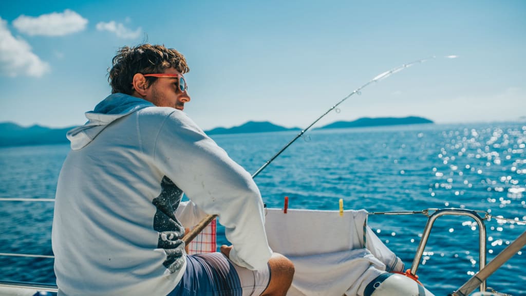 A person on a boat, wearing sunglasses and a hoodie, holds a fishing rod over the blue ocean near Playa del Coco. With majestic mountains and a clear sky as their backdrop, they're enjoying the adventure of fishing in Costa Rica's waters.